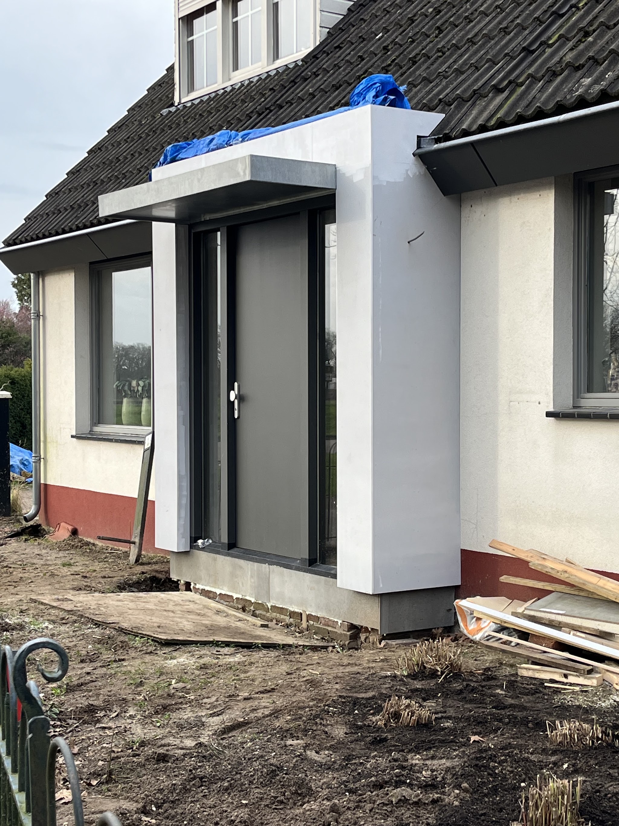 Nieuwe entree met grijze voordeur en wit gestucte aanbouw, de tuin nog in aanleg
