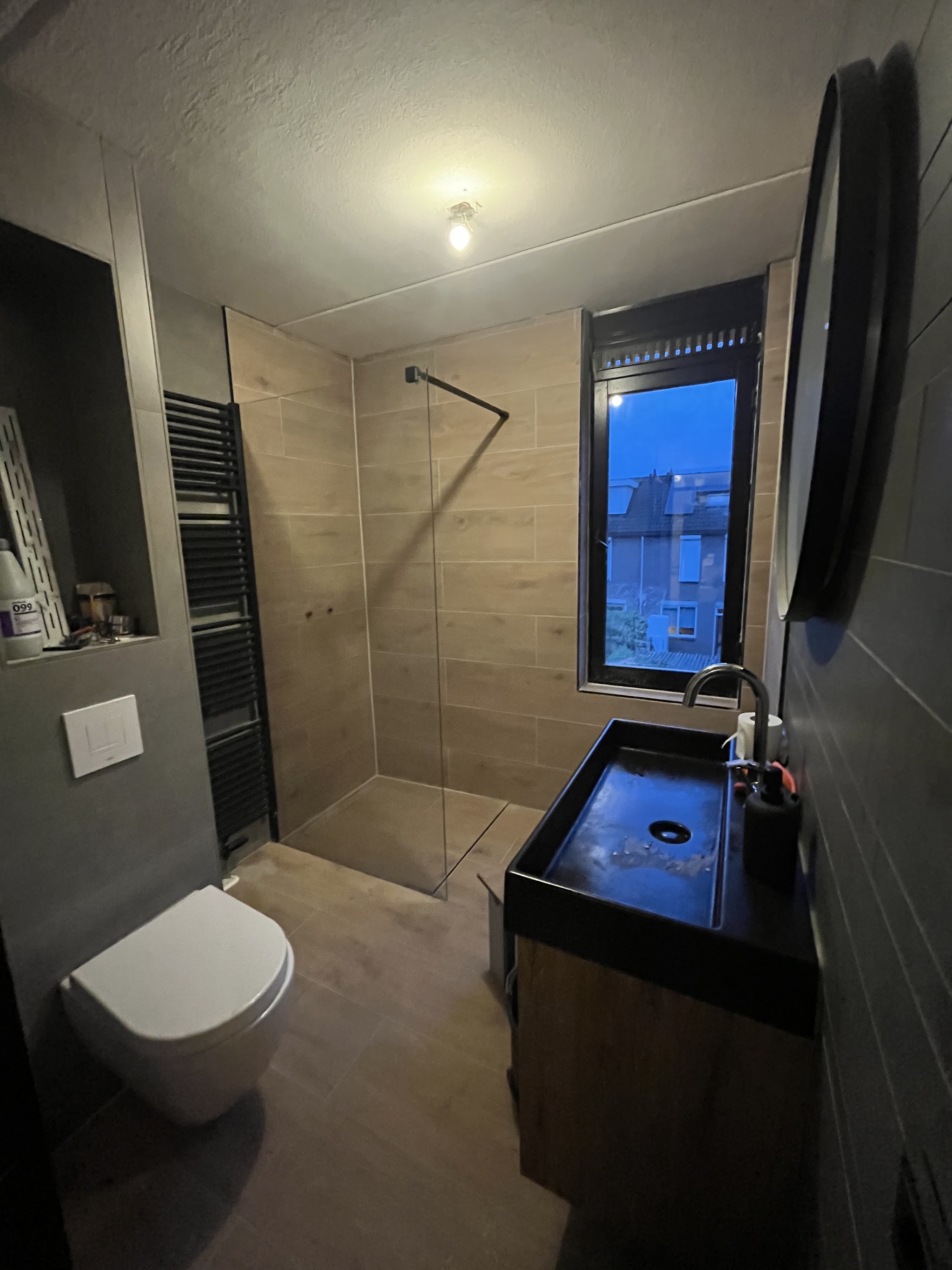 Afgewerkte badkamer met inloopdouche, houtlook tegels, zwarte wastafel op een houten meubel en een designradiator